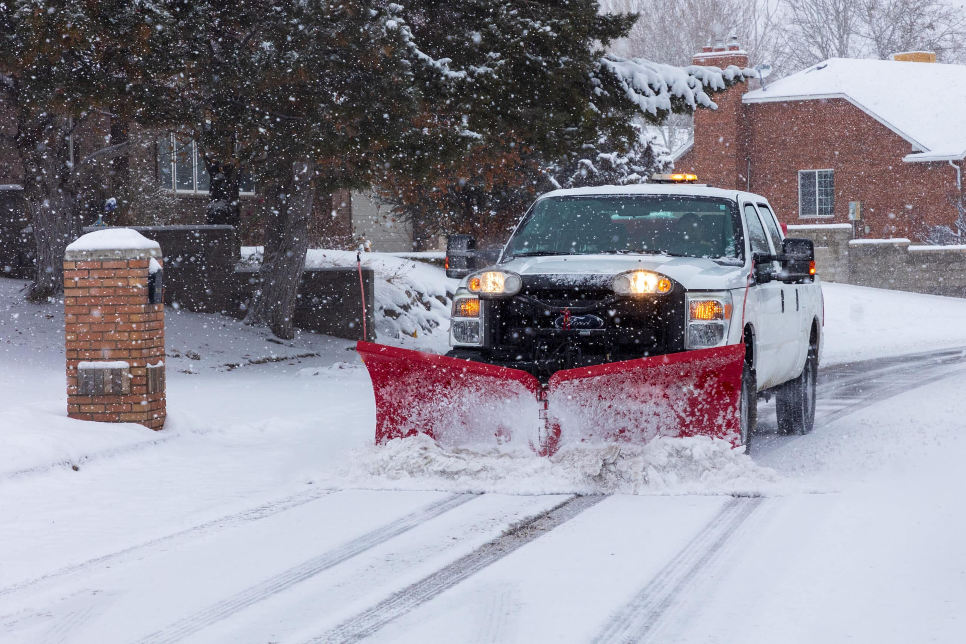 Snowplow truck with a red blade clearing a residential street during snowfall
